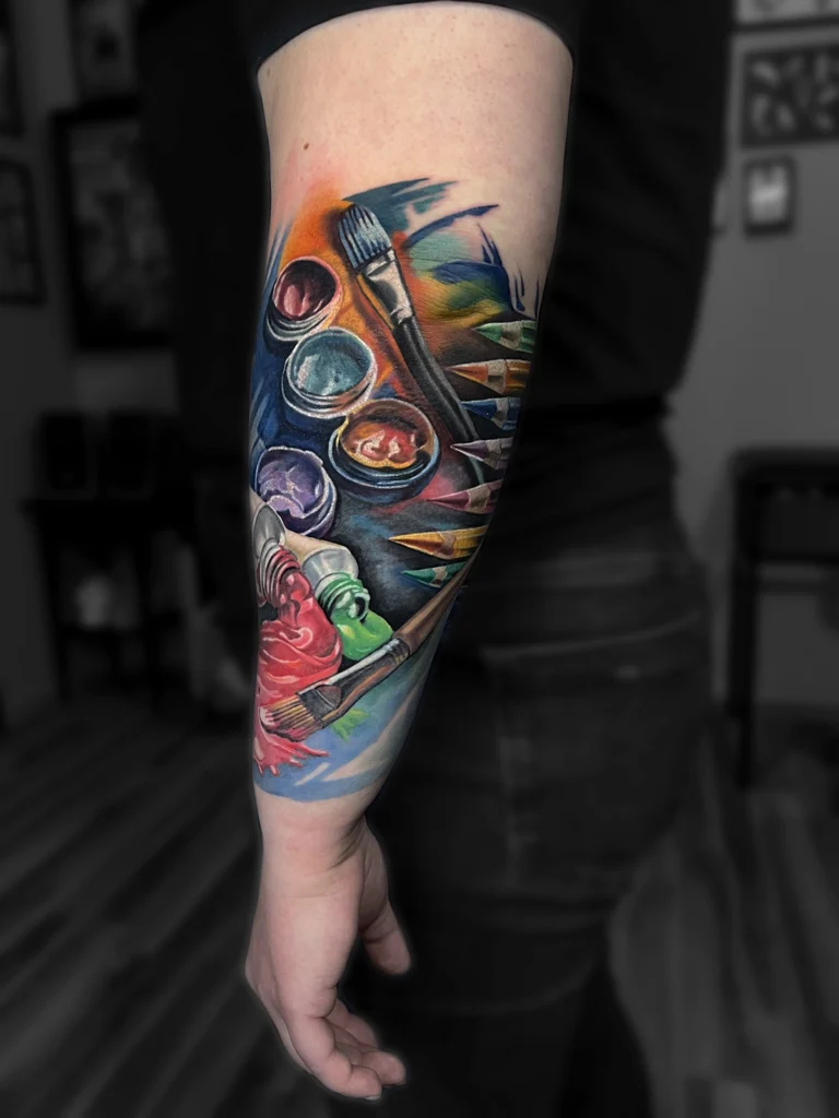 Kolorowy realistyczny tatuaż przedstawiający pędzle, farby i przybory malarskie wykonany przez Bartka w Studio Fortuna Tattoo w Warszawie.